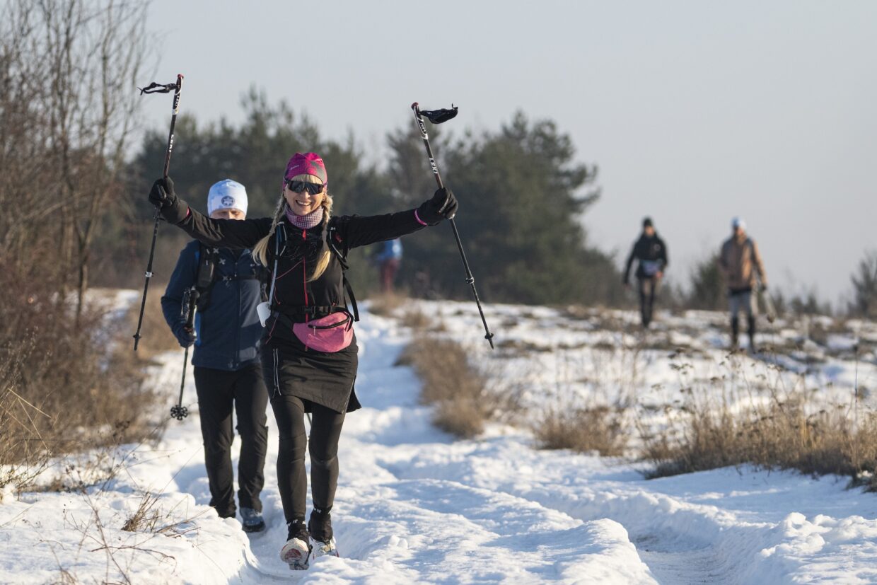 Uśmiechnięta uczestnicza maratonu podnosi kijki trekkingowe w górę. W tle widać innych uczestników.